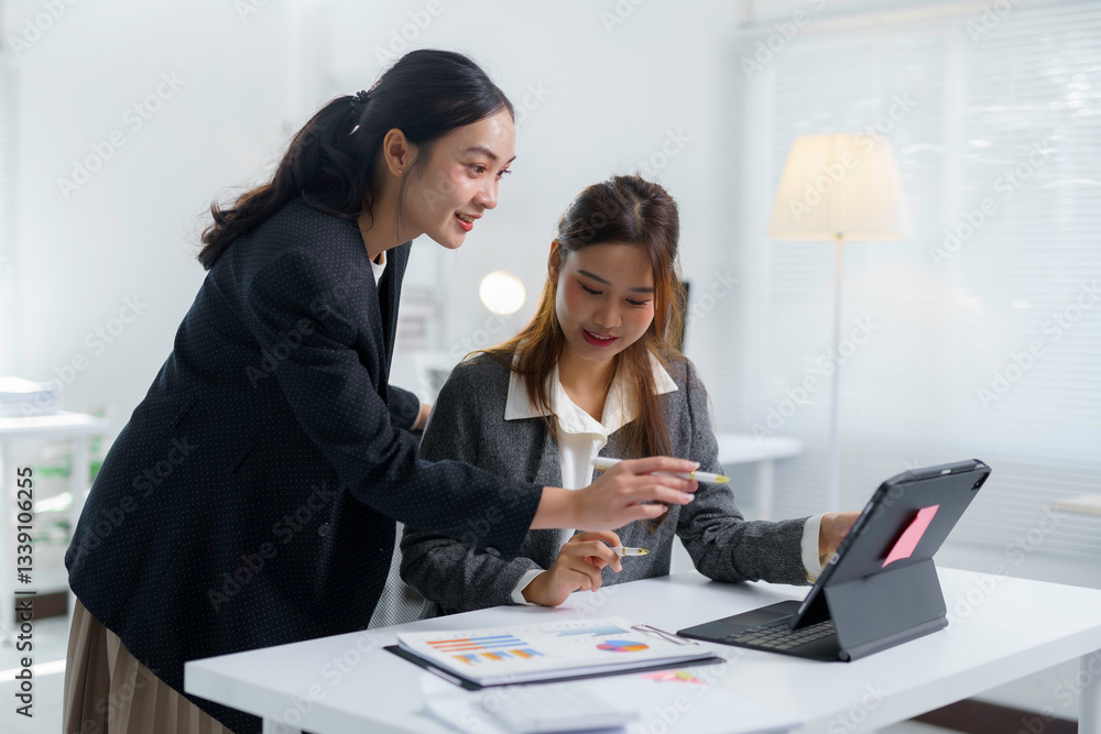 Poster two asian businesswomen discussing work while utilizing a tablet and reviewing documents in a modern