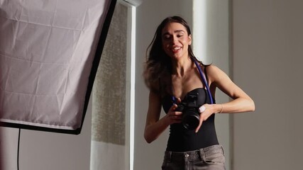 A young woman photographer sets up lighting equipment in a spacious studio. She wears a black tank top and has a camera hung around her neck, getting ready for an upcoming shoot.
