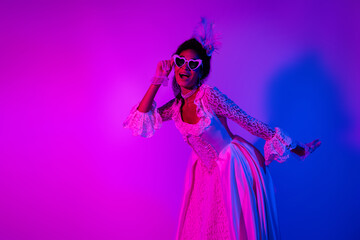 Young woman in a glamorous lace gown with heart-shaped glasses amidst vibrant pink and purple neon lights.