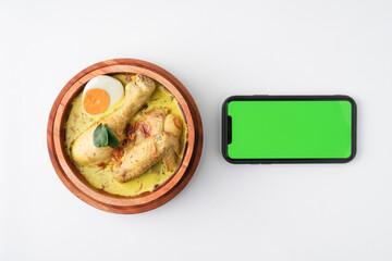 Blank green screen smartphone and Lontong opor ayam served during eid Mubarak, Eid al Fitr holiday celebration with Ketupat Lebaran or rice cake dumpling and sambal chili sauce.