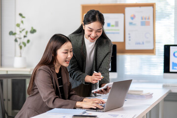 Two women are sitting at a desk with a laptop and a stack of papers