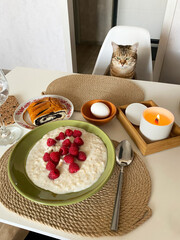 Curious Cat sits on a chair at the kitchen table set with a breakfast of porridge and berries, an egg and a candle burning for atmosphere. Cat at breakfast