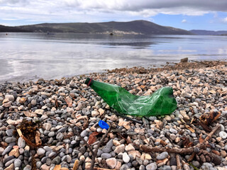 Crumpled plastic water bottle and cap lying on public pebble beach, trash on beach, view of sea bay