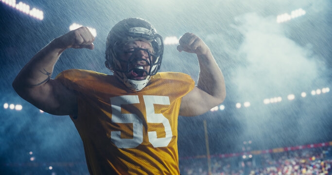 Portrait of an American Football Player Celebrating a Successful Game Victory and Raising Arms in Triumph. Male Athlete in Yellow Jersey, Helmet and Gloves Showing Confidence, Flexing Muscles