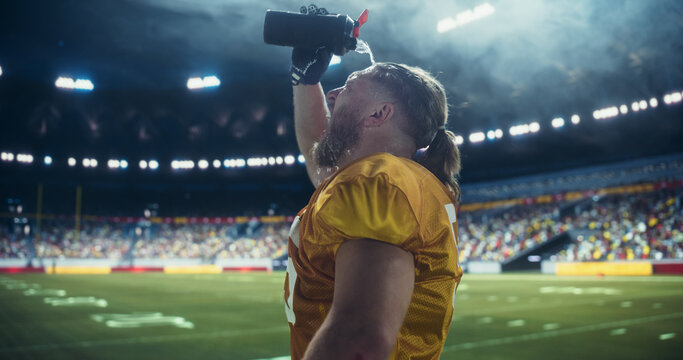 American Football Player Pouring Water on His Face During Championship Game in Stadium. Male Athlete Hydrating on Field with Crowd in Background. Professional Sportsmen Drinking Water - Powered by Adobe