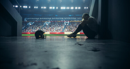 American Football Player Sitting Alone in a Stadium Tunnel, Tossing Away His Helmet. Background with a Crowded Stadium with Sport Fans. Footballer is Emotional Devastated After a Championship Game