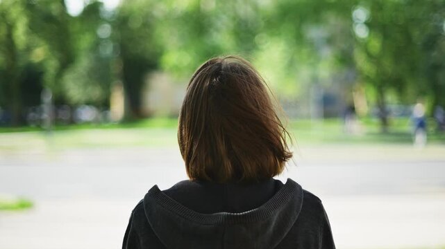 Rear view of adult brunette female standing on the street and looking at traffic and buses passing by, 4K