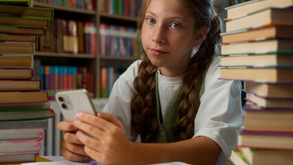 Bored calm relaxed Caucasian little girl kid schoolgirl school child pupil student learner sitting desk between two piles of books use mobile phone online app playing game smartphone in school library