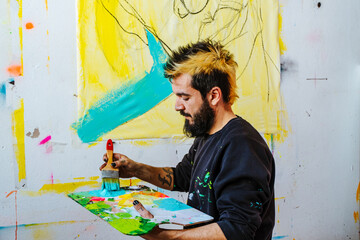 Artist Painting an Abstract Canvas with Blue and Yellow. An Argentine artist applies blue paint to a yellow-toned canvas, working on an expressive abstract piece in his creative studio.