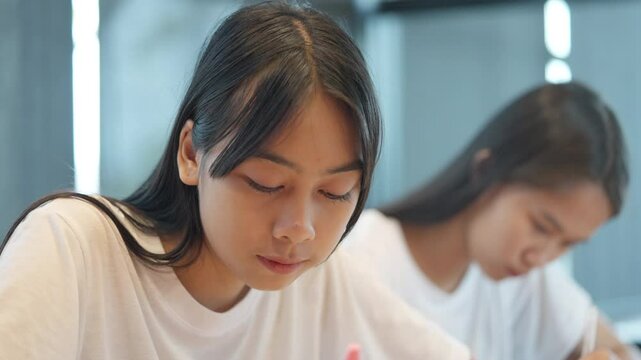 Two girls are sitting at a desk, one of them is looking at a book. The other girl is looking at her own book. Scene is focused and studious