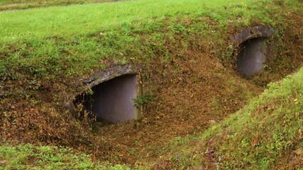 Close-up shot of a grassy field with two concrete bunkers in the middle. A couple of bunker entrances on a grassy knoll, on a historical battlefield site, in the rain.