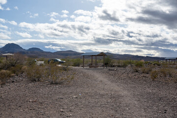 Dieses Bild zeigt eine Au&szlig;enszene mit einem gepflasterten Weg, der zu einer Holzstruktur f&uuml;hrt, die wie ein Park oder Picknickplatz aussieht und sich in einer w&uuml;sten&auml;hnlichen Umgebung befindet. 