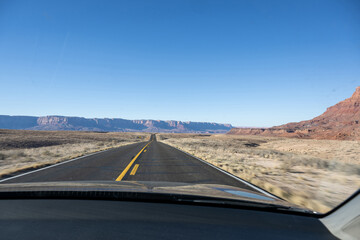 Eine offene Stra&szlig;e in der W&uuml;ste, die zu ikonischen roten Felsformationen vor strahlend blauem Himmel f&uuml;hrt.
