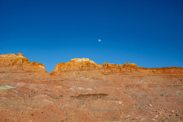 Das Bild zeigt eine weite W&uuml;stenlandschaft unter strahlend blauem Himmel, mit schroffen Bergen und Schluchten im Hintergrund und einem Vollmond, der &uuml;ber dem Horizont aufgeht.