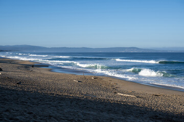 ine ruhige Strandszene mit einem Sandstrand, der unter blauem Himmel sanft ins Meer abf&auml;llt. Schaum von Wellen bildet eine Kulisse f&uuml;r den Vordergrund. In der Ferne sind einige M&ouml;wen zu sehen