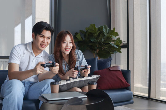 Excited young Asian couple holding game controllers to play game together during sitting on sofa at living room. Concentrated two close friends holding joystick to play video game online at home.