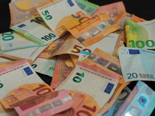 Different denominations of euro bills are stacked in a chaotic order, creating a three-dimensional effect. The black background enhances the contrast