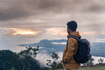A traveler stands on a mountain peak at sunrise, gazing at the misty valley and ocean. Wearing a...
