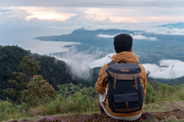 A traveler sits on a mountain peak, gazing at the misty valley and ocean at sunrise. Wearing a...