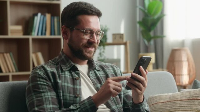 Smiling happy relaxed middle-aged man at home living room holding cell mobile phone using smartphone on couch online scrolling social media male guy checking apps buying shopping texting messages