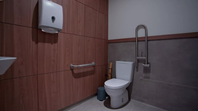 A modern accessible restroom with a white toilet, stainless steel grab bars, a wall-mounted paper dispenser, and wooden wall panels, adjacent to a sleek hallway with bright lighting.