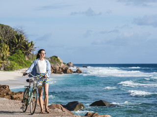 Cute woman with vintage bicycle on the background of stunning beach with palm trees and rocks. Beautiful views and sights of Seychelles. Clear, sunny day. Concept of leisure and travel