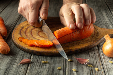 A person skillfully cuts bright orange carrots on a rustic wooden board, surrounded by garlic and...