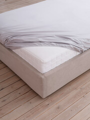 Bed in a bright bedroom with wooden floors is covered with grey fitted sheet with an elastic band, but the corner of the white mattress is visible. Side view photo. Suitable for mock-up.