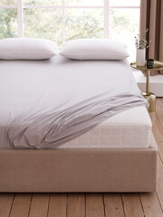 Bedroom with window, wooden floor, double bed with mattress, covered by fitted sheet and two pillows. Mattress corner is visible. Bedside table next to the bed. Front view photo. Suitable for mockup