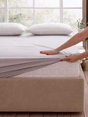 Woman is putting a sheet on a bed. One corner of the mattress is visible. The bed with two pillows is on the wooden floor of bright modern bedroom with a big window. Front top view photo