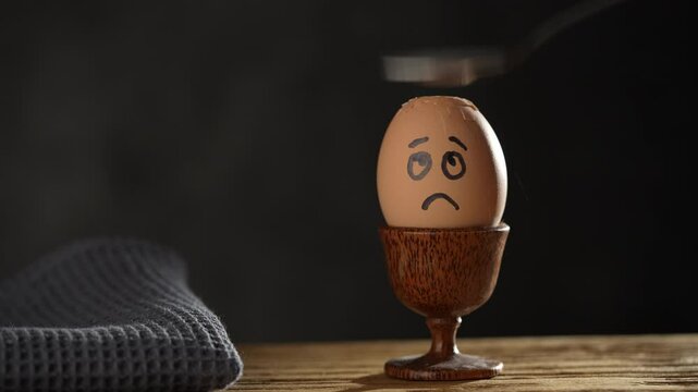 Metal spoon breaking boiled egg with sad face image in a eggcup on a wooden table, close up. Scared egg looking at spoon