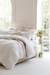 Sunny bedroom with two large windows. Four pillows, cushion and a fluffy down comforter on a double bed with bedside table and bouquet in vase on it. Side view close-up photo. Suitable for mockup