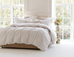 Sunny bedroom with two large windows. Four pillows, cushion and a fluffy down comforter on a double bed with bedside table and bouquet in vase on it. Side view horizontal photo. Suitable for mockup
