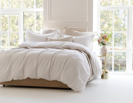 Sunny bedroom with two large windows. Four pillows, cushion and a fluffy down comforter on a double bed with bedside table and bouquet in vase on it. Side view horizontal photo. Suitable for mockup