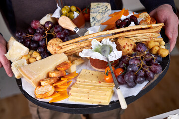 A man holds a wooden Charcuterie Board with snacks, cheese, sausages and fruit, close up