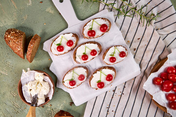 Creative tartines with white cheese and tomato cherries rest on a white board on a green table and a striped napkin