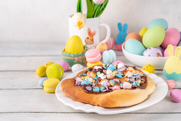 Sweet Easter dessert pizza, with chocolate paste topping, marshmallow and Easter chocolate eggs, snack for Easter holiday kids party, with festive decorations, colorful eggs and bunny rabbits