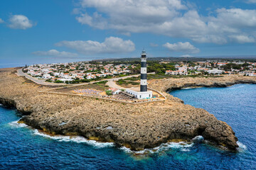 Obraz premium Artrutx Lighthouse perched on rocky coastline, black-white stripes contrasting turquoise Mediterranean waters near coastal Menorca settlement, Balearic Islands landscape