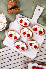 Creative tartines with white cheese and tomato cherries rest on a white board on a green table and a striped napkin