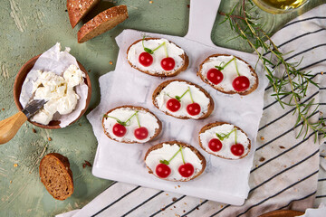 Creative tartines with white cheese and tomato cherries rest on a white board on a green table and a striped napkin