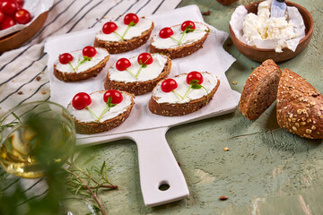 Creative tartines with white cheese and tomato cherries rest on a white board on a green table and a striped napkin