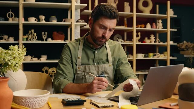 Thoughtful focused Caucasian man male guy small business owner potter salesman businessman craftsman thinking counting income debt finance budget use laptop calculating salary bill calculator indoors
