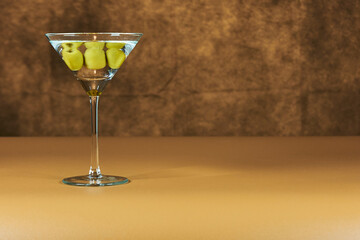 Refreshing Martini glass with green olives inside, on a warm colored surface.