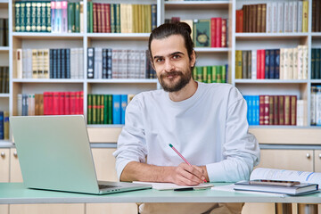 Young male teacher working in library sitting at desk with laptop books, looking at camera