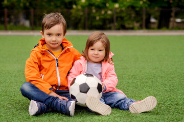 Fototapeta premium Two children, boy and girl, in bright jackets sitting on green football field holding soccer ball. Kids, brother and sister, watching game. Competition, active childhood, and team support concept