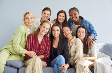 Fototapeta premium Portrait of happy young diverse women group looking cheerful at camera sitting on sofa on gray wall background. Female business people or company employees in casual clothes smiling indoors.