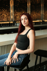 a red head ginger brunette model young woman teenager sits at a bar with gold lights and black designs while wearing a tanktop and jeans