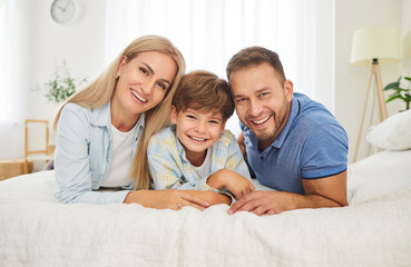 Obraz premium Joyful portrait of parents embracing son on a bed at home bedroom and looking at camera. Smiling and bonding, family love, togetherness, and a happy moment in a relaxed atmosphere.