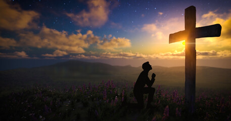 Silhouette of a man praying in front of the cross on a meadow with moving clouds