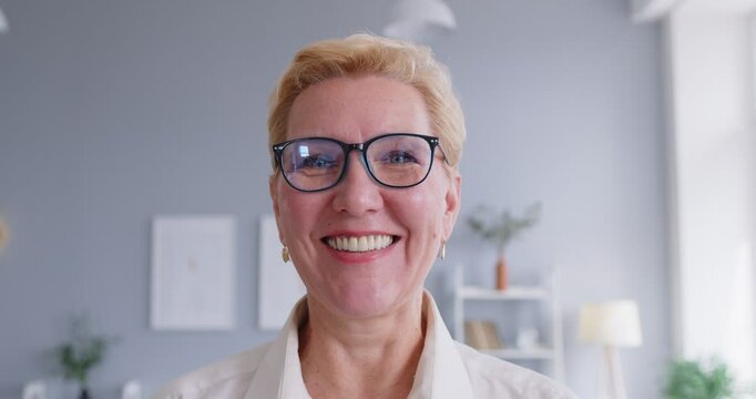 Happy middle aged woman puts on glasses. Head shot video portrait of cheerful beautiful short haired mature woman who puts on her eyeglasses, smiles and looks at camera